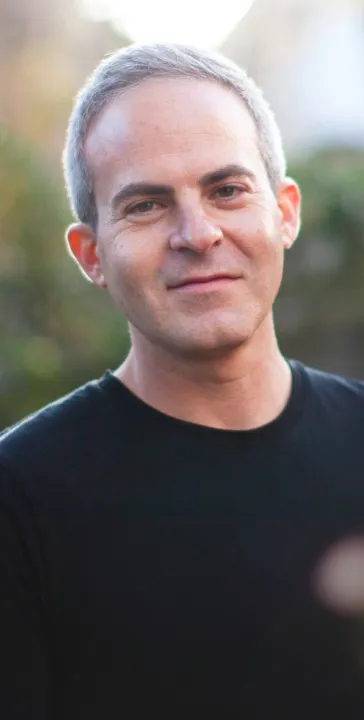 Portrait of a smiling middle-aged man with short gray hair wearing a black shirt against a blurred outdoor background.